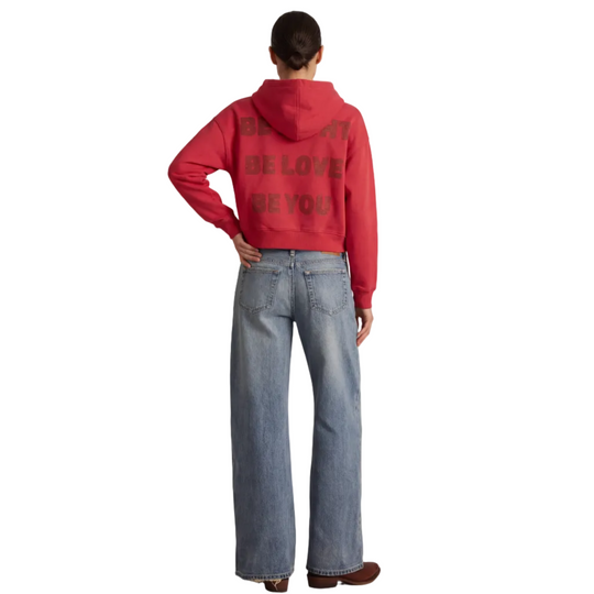 Person wearing a red hoodie with text on the back and blue jeans, standing against a white background
